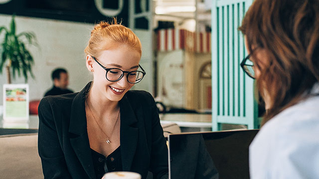 Women looking over the laptop and talking to another women in front.