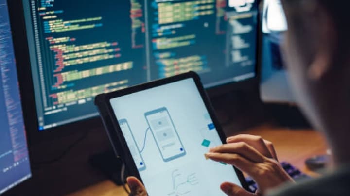 Developer designing a mobile app on a tablet with code on screens in the background