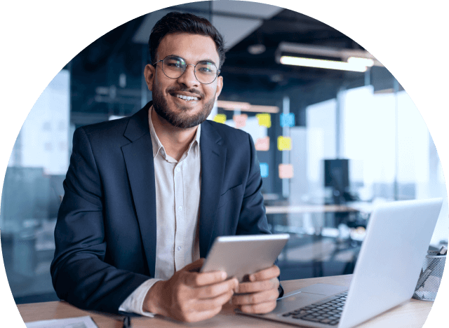 A person smiling at a desk with a tablet and laptop in an office.